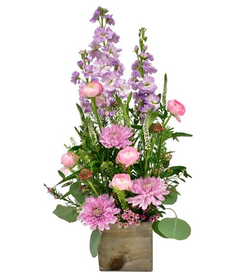 A tall floral arrangement featuring pink ranunculus, lavender stock, and mums, accented with lush greenery, displayed in a rustic wooden container.